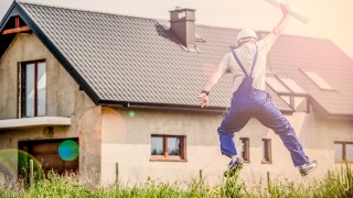 Handwerker im Blaumann springt freudig vor einem Haus